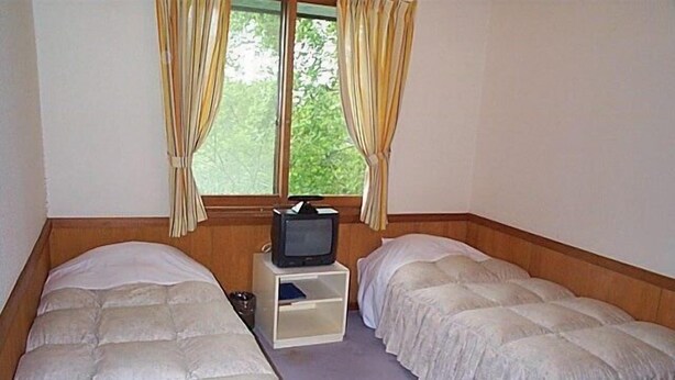 A small, simple room with two single beds, a television on a small table, and a window with curtains. - Pension Noichigo (Suzaka)