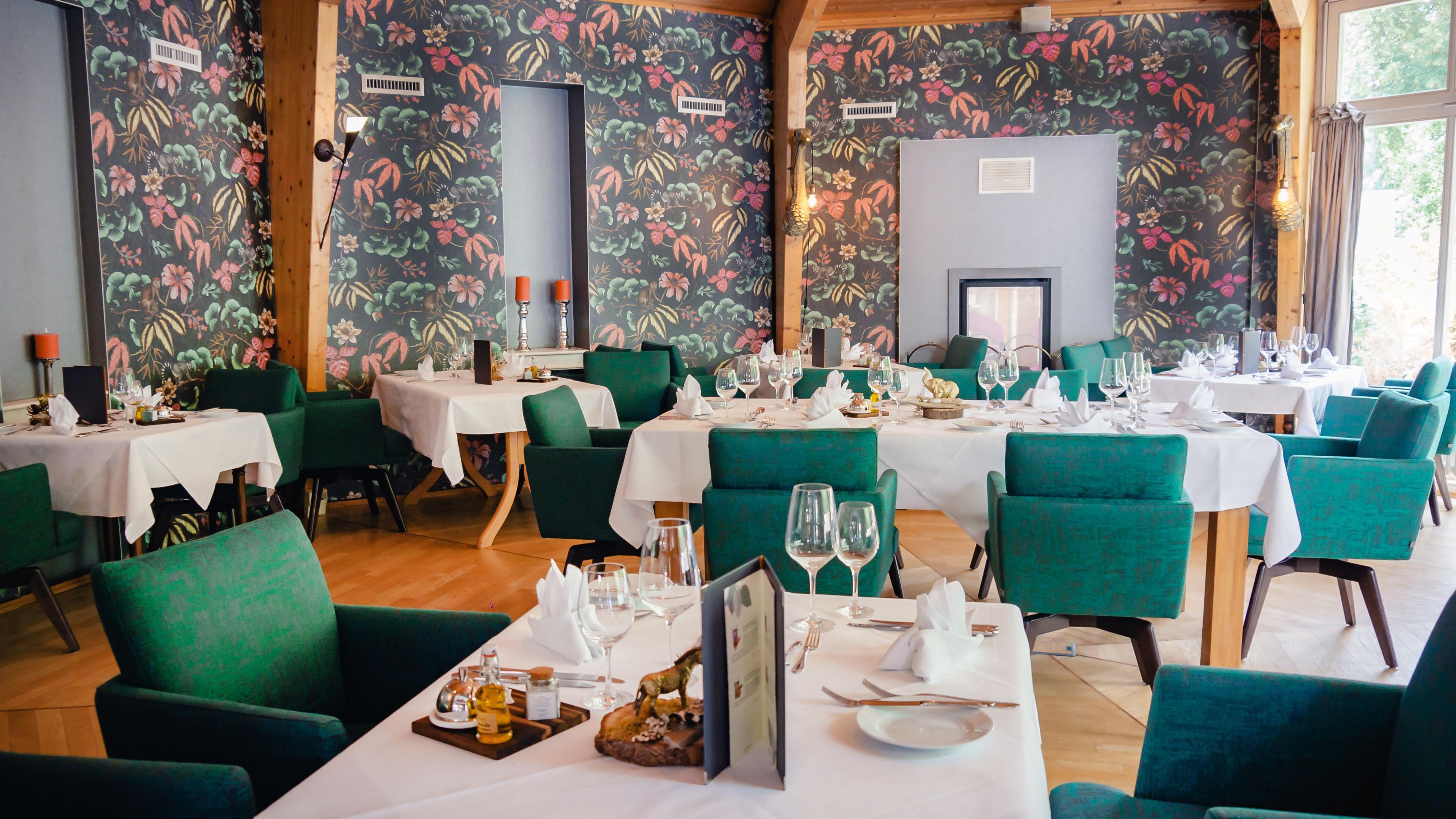 A restaurant with green upholstered chairs, white tablecloths, and floral wallpaper.