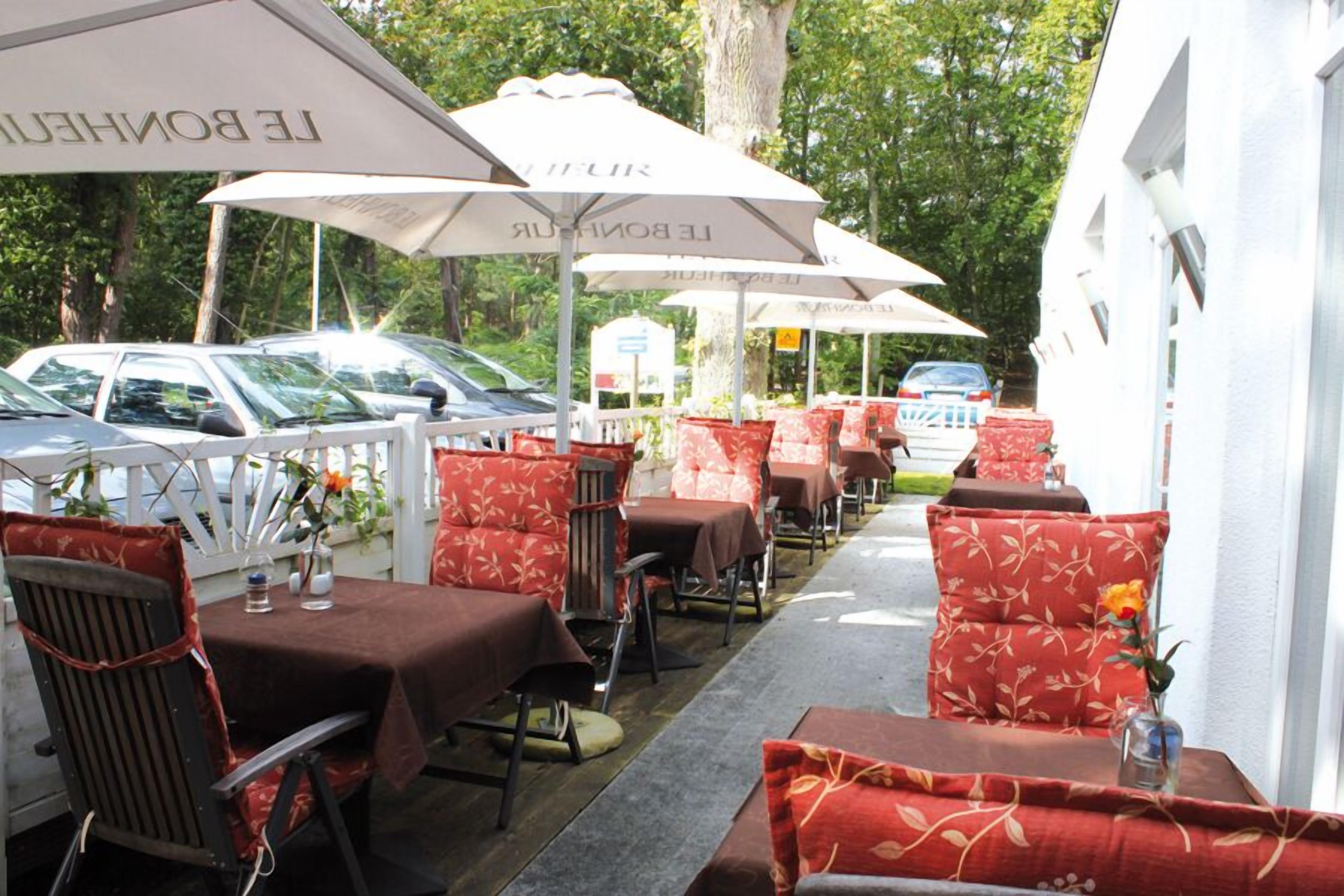 Un espace extérieur aménagé pour les repas, avec des tables, des chaises, des parasols et une rangée de voitures garées.