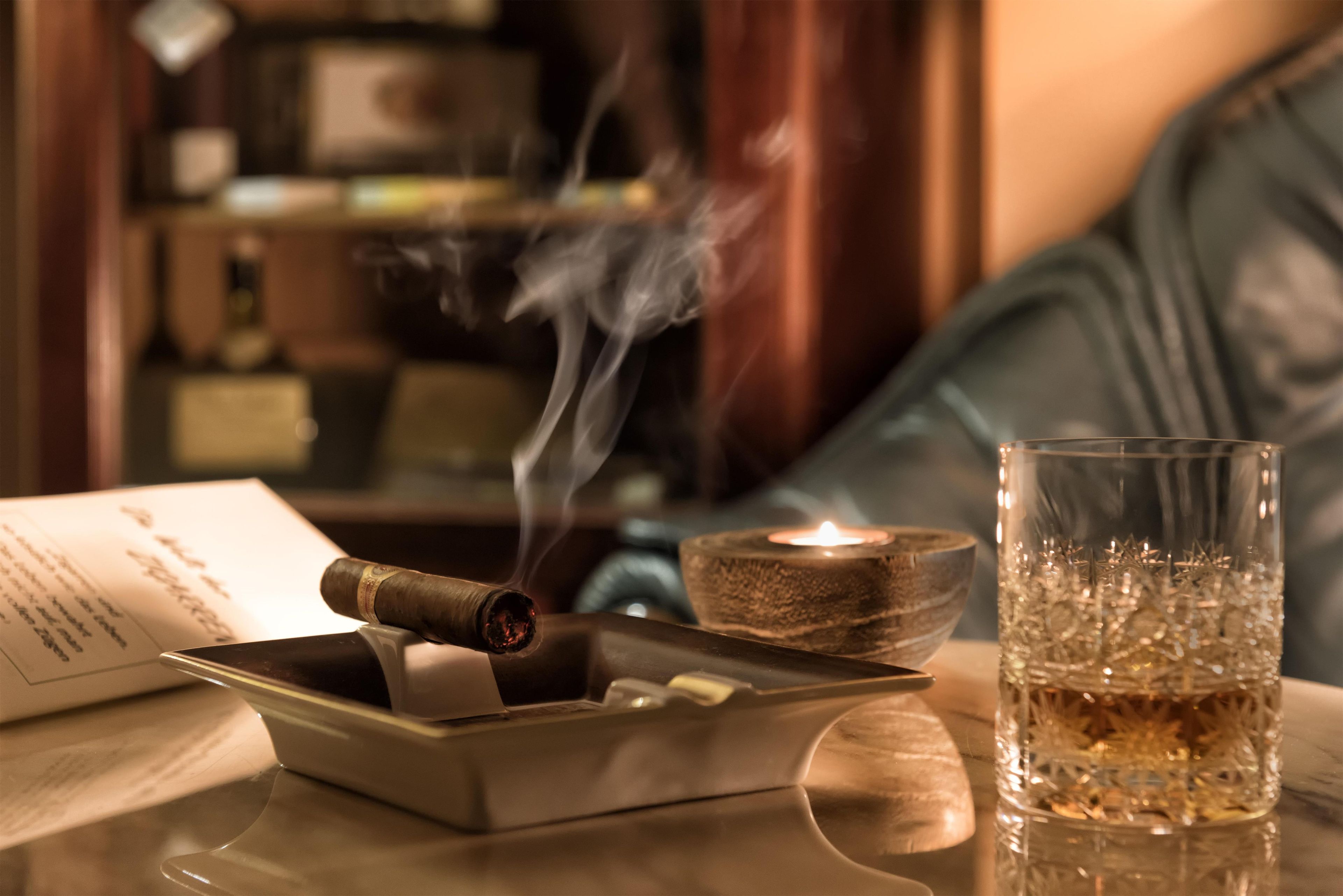 A cigar in a ashtray, a lit candle, and a glass of whiskey on a table.