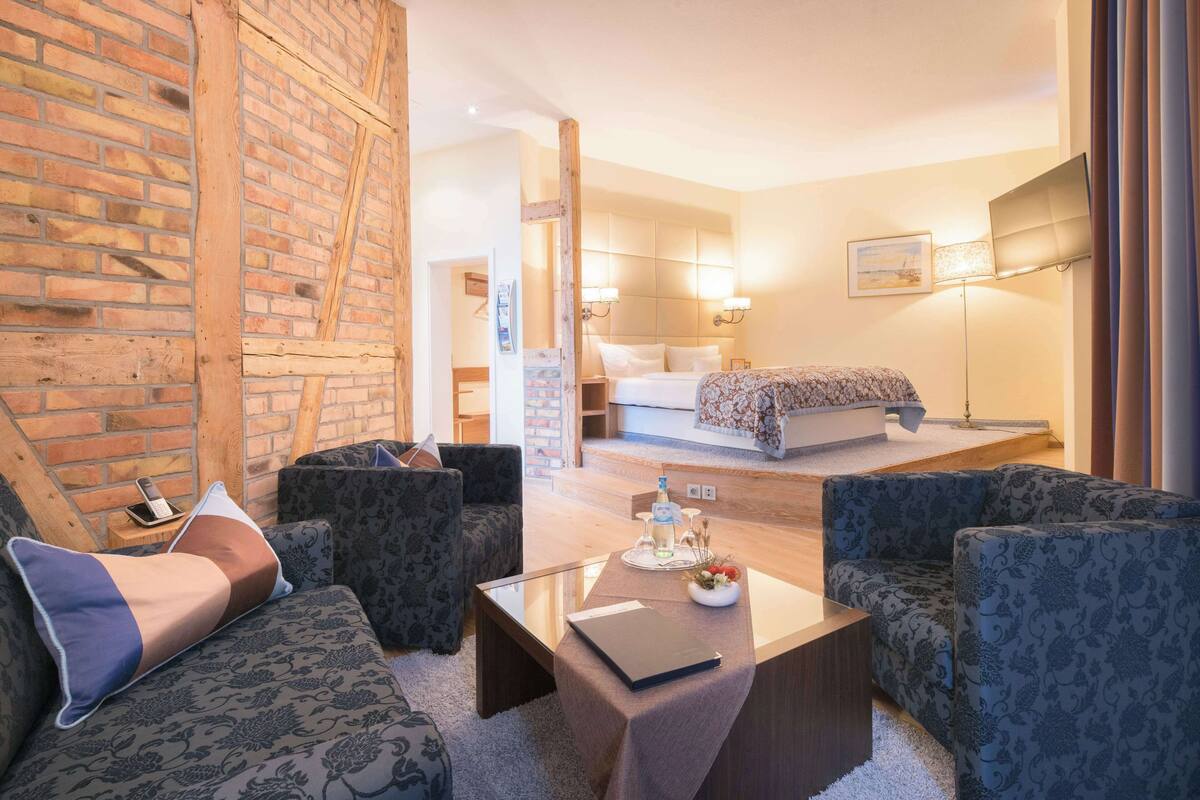 Een moderne hotelkamer met een bakstenen muur, een bed met een gestreept beddegoed, een zithoek met beklede banken, een glazen salontafel en een nachtkastje met een lamp en een fles.