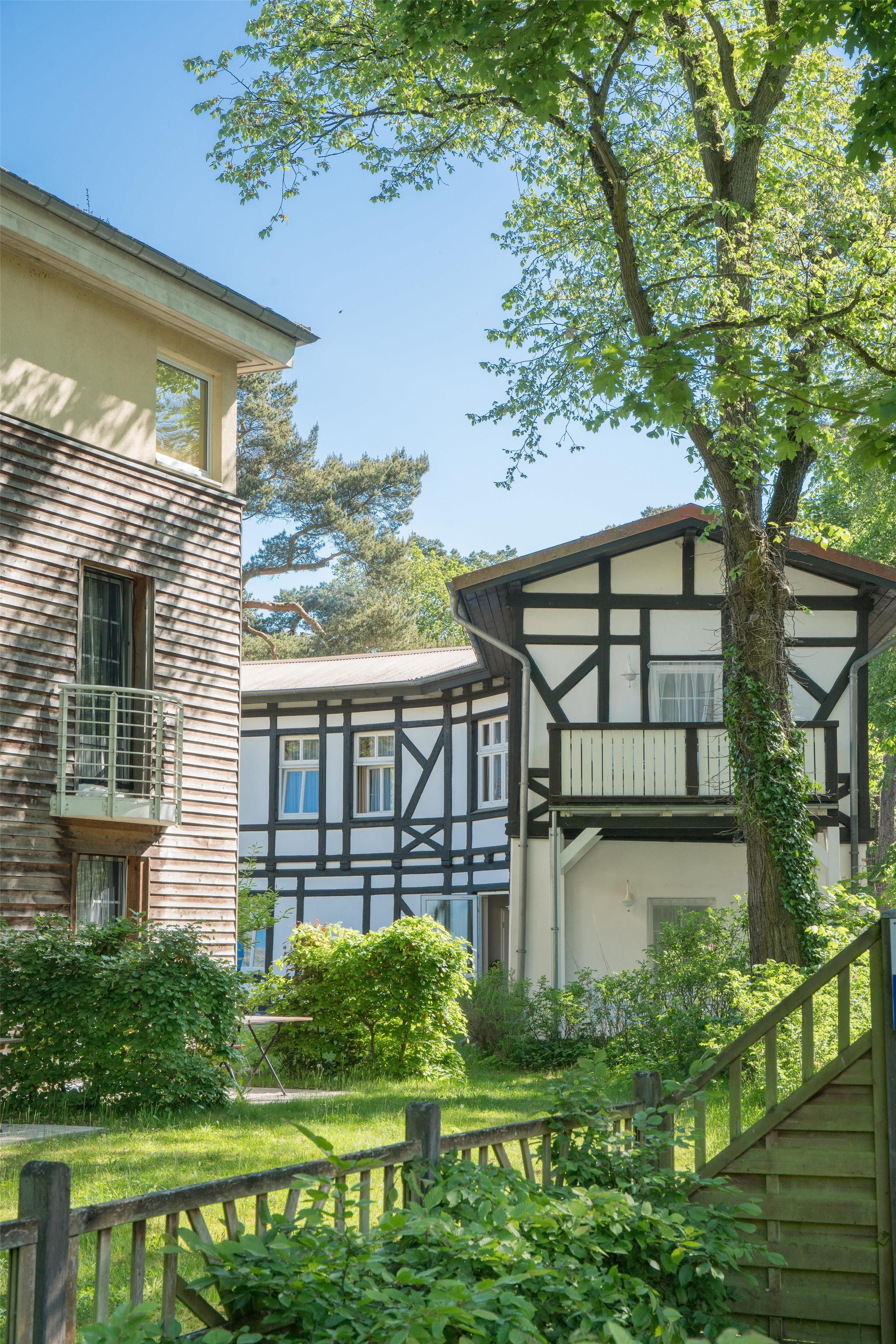 A traditional half-timbered house with a balcony, surrounded by lush greenery and a wooden fence.