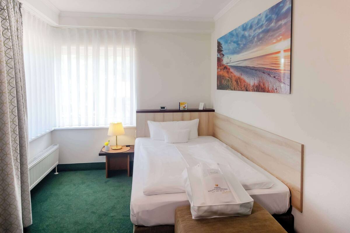 Een hotelkamer met een bed, een nachtkastje met lamp, een schilderij aan de muur en een raam met gordijnen.
