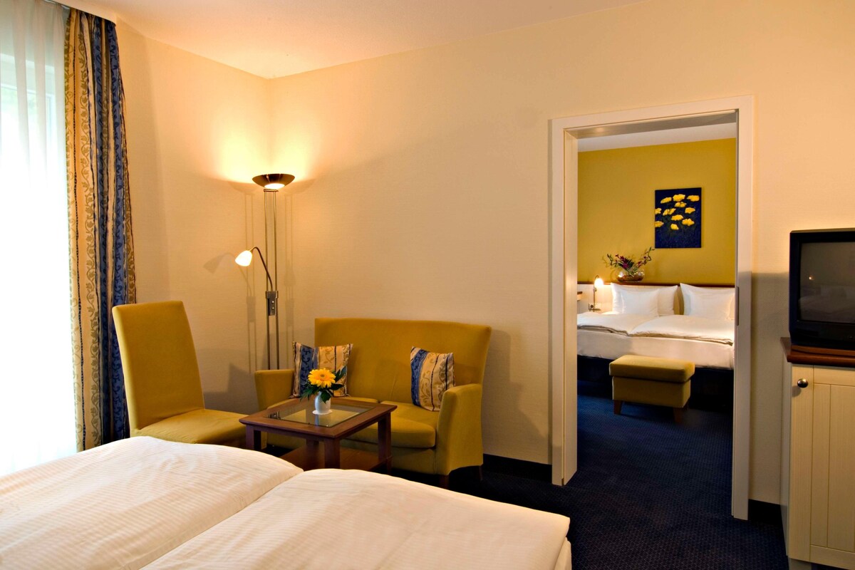 A hotel room with two beds, a sofa, a small table with a vase, a lamp, and a television.