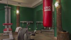 Une salle de sport aux murs verts, au sol en bois et équipée de matériel d’exercice, notamment un sac de frappe rouge sur lequel est inscrit « U.N.O. SPORTS ».