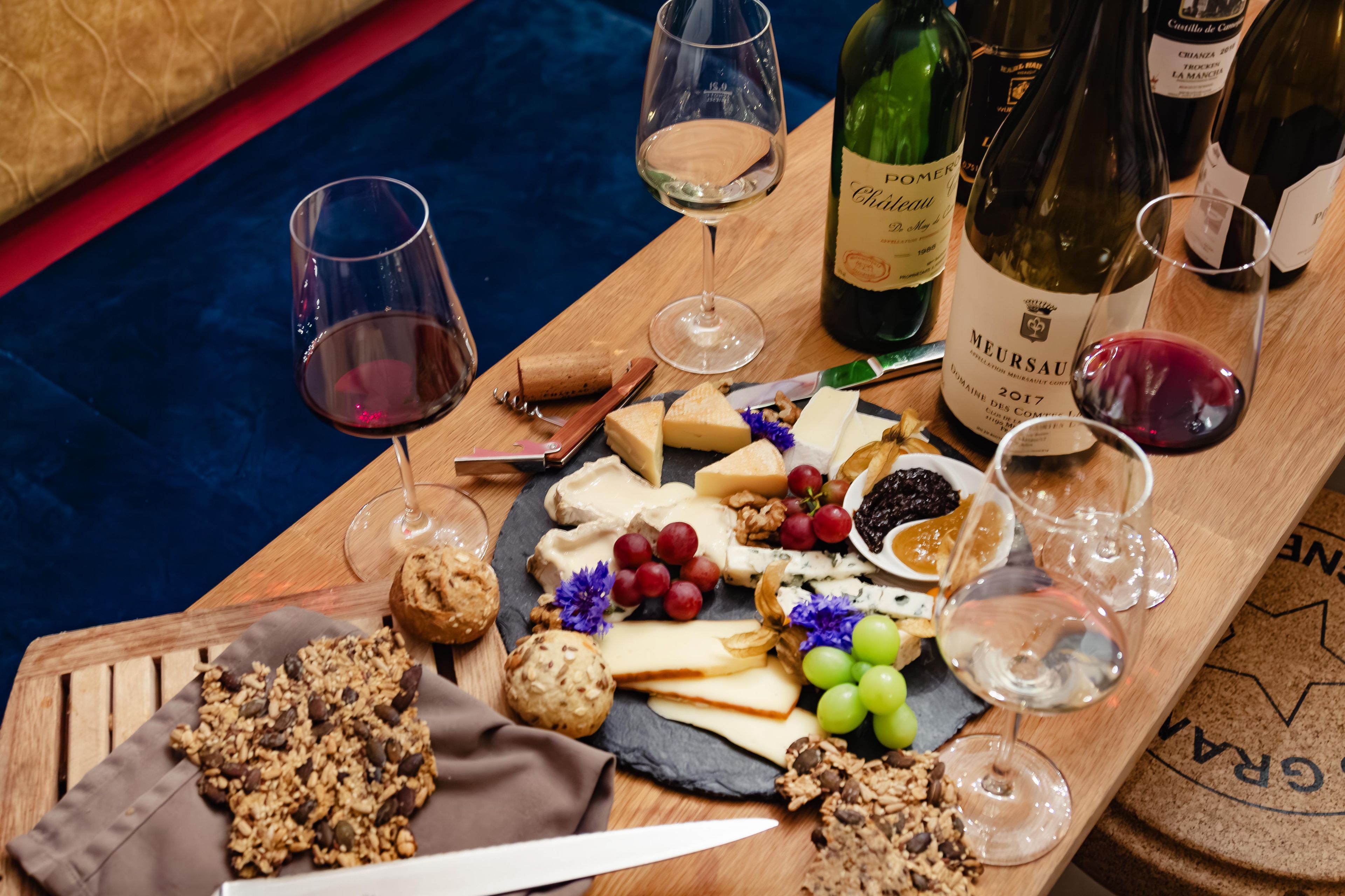 Una tabla de madera con una variedad de quesos, frutas, nueces y galletas, acompañada de copas y botellas de vino.