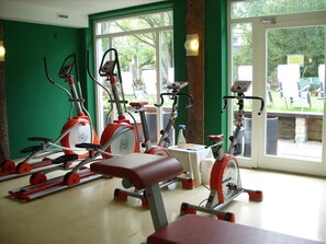 Een sportschool met hometrainers en een bank, grote ramen en een groene wand.
