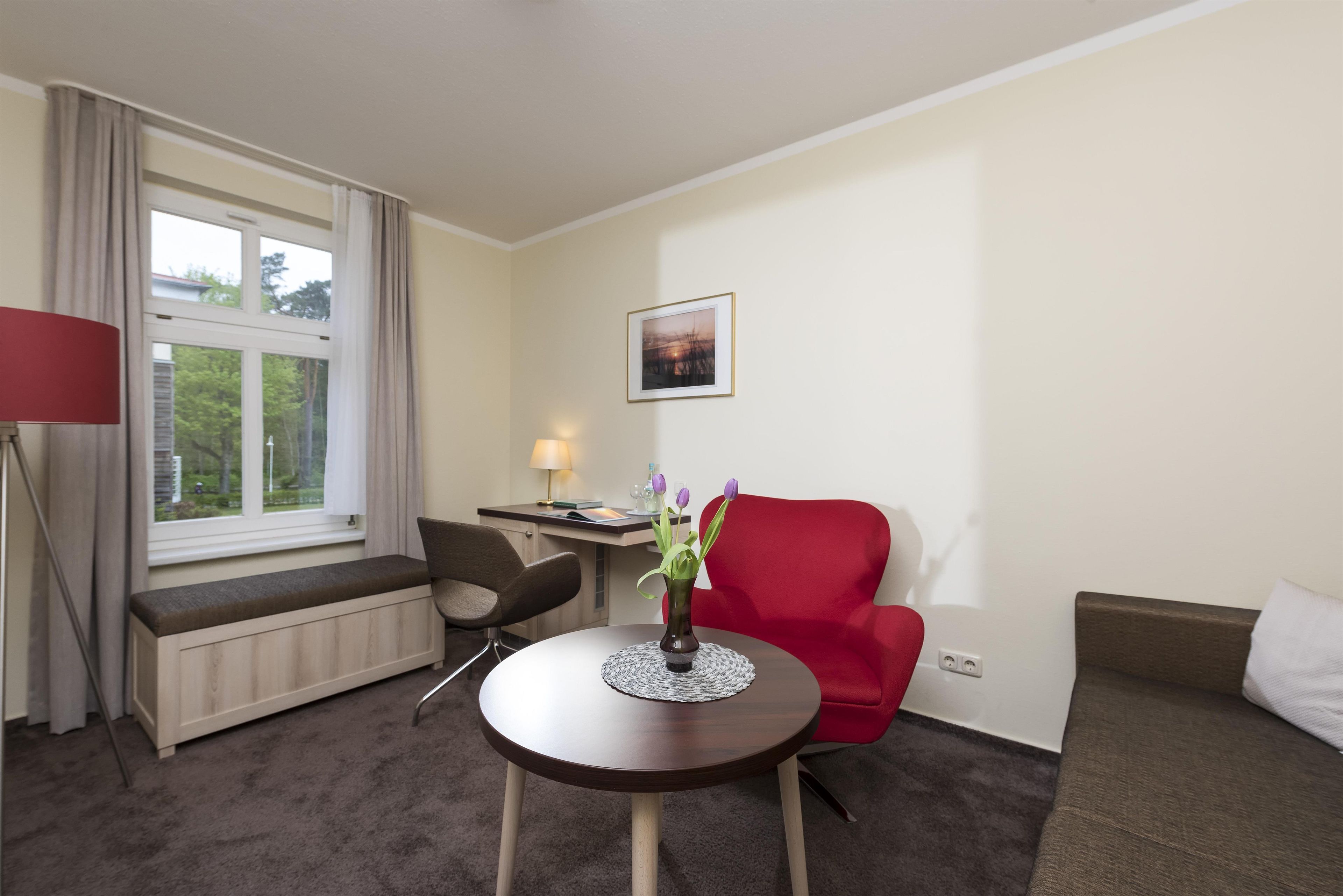 Une chambre d’hôtel avec une table ronde, une chaise rouge, un bureau avec une lampe, une fenêtre avec des rideaux et un tableau encadré au mur.