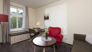 Une chambre d’hôtel avec une table ronde, une chaise rouge, un bureau avec une lampe, une fenêtre avec des rideaux et un tableau encadré au mur.