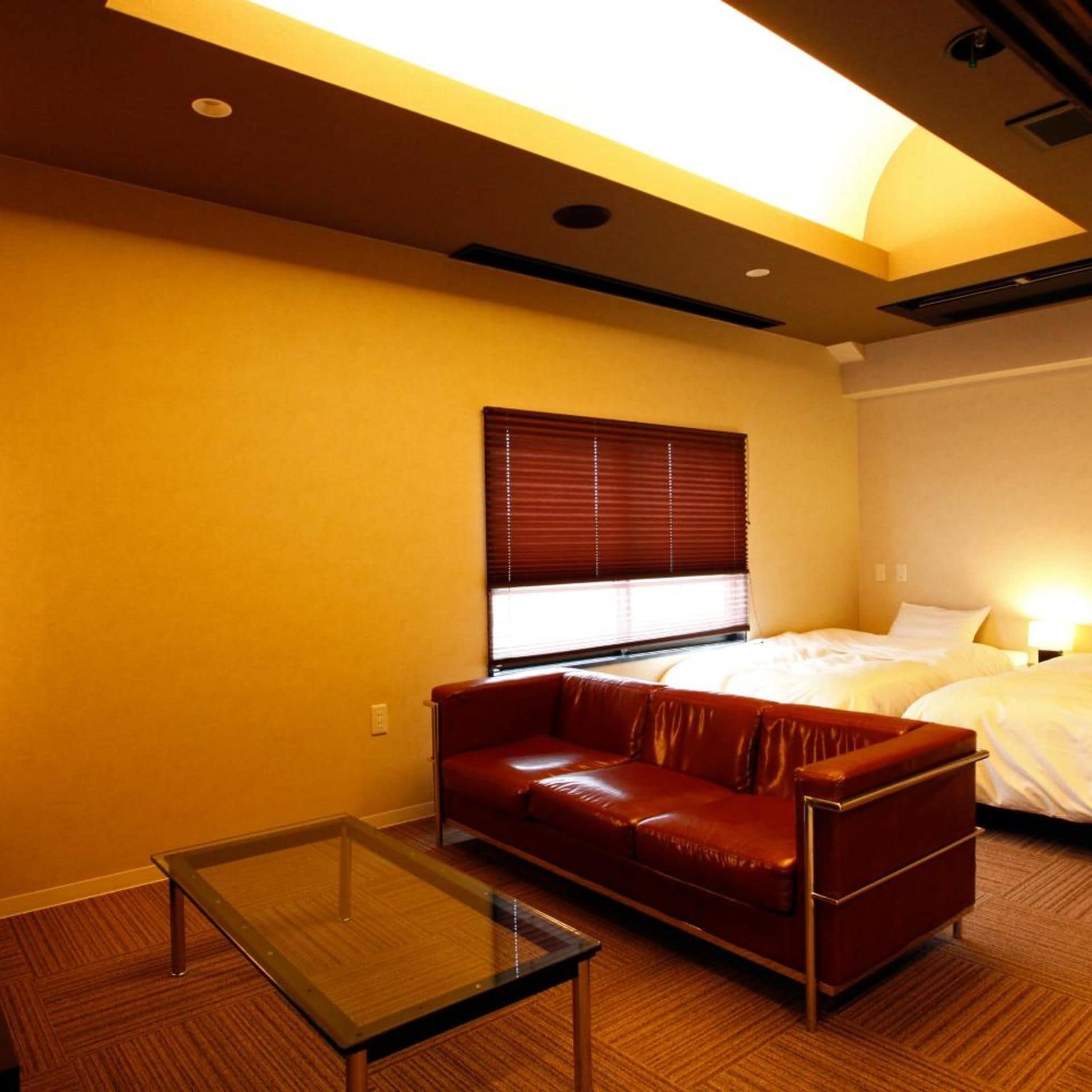 Quarto de hotel com sofá de couro, mesa com tampo de vidro, cama com roupa de cama branca e janela com persianas.