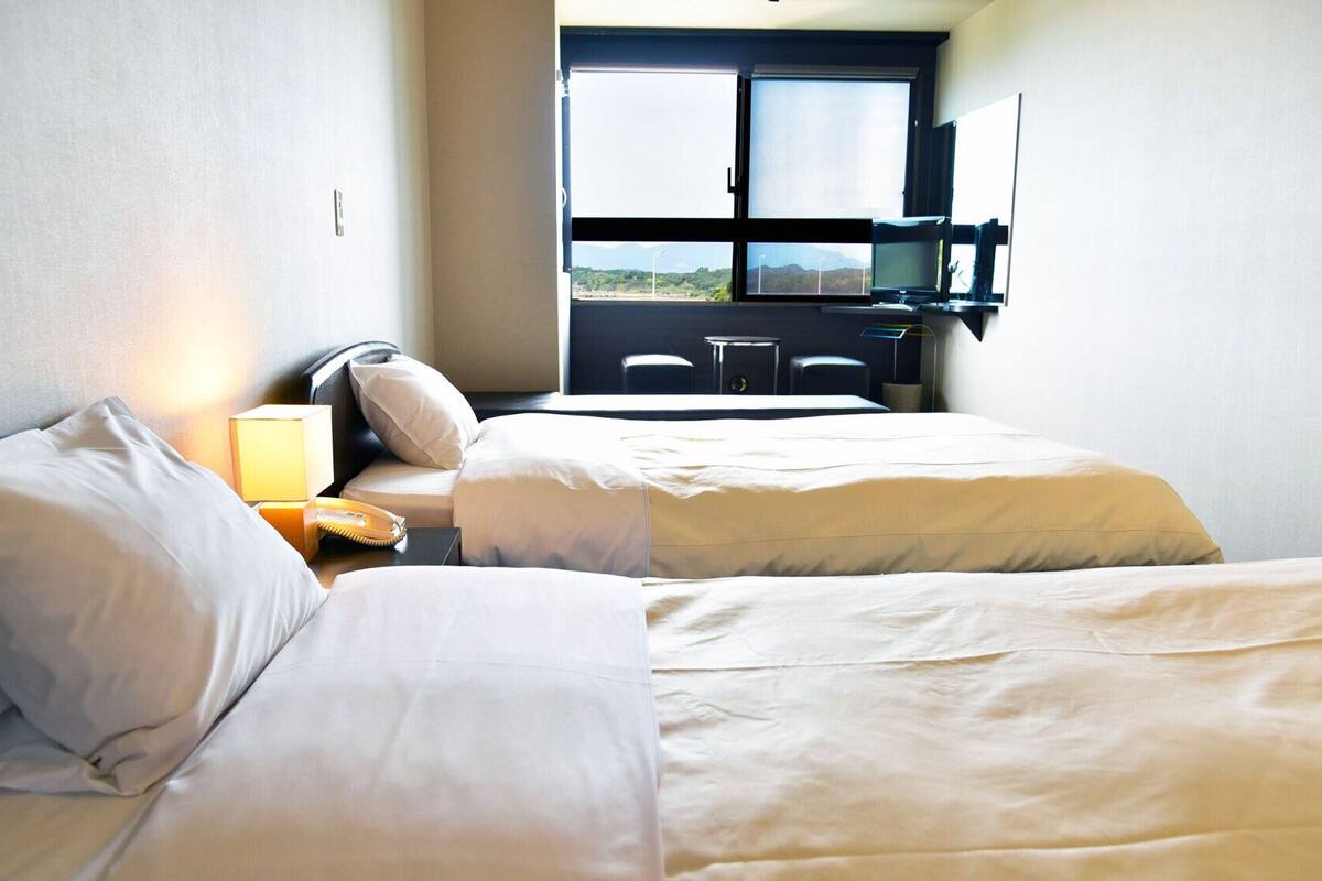 A hotel room with two beds, a desk, a chair, a lamp, and a window with a view.