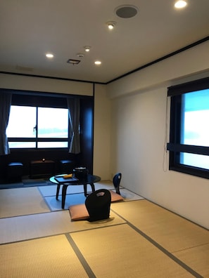 Ein Zimmer mit Tatami-Matten, einem niedrigen Tisch mit Stuhl, einer Couch und einem großen Fenster mit Vorhängen.