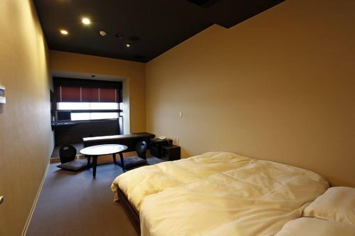 A bedroom with a bed, a small table, and a chair. There is a window with blinds and a door on the left.