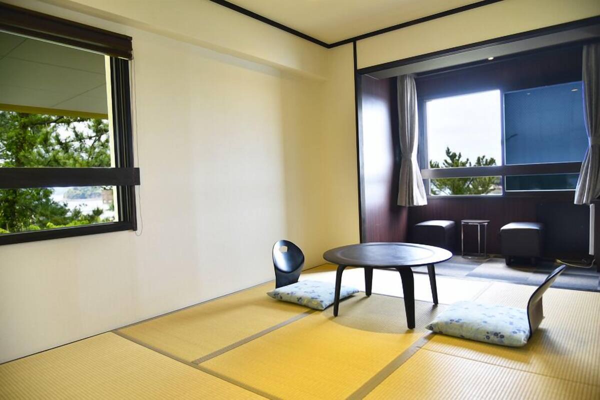 A room with tatami flooring, a round table, two cushions, a chair, and a large window with curtains.