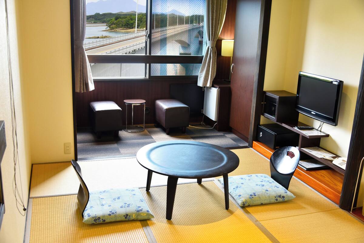 A room with tatami flooring, a tatami mat table, two cushions, a television, and a view of a bridge and greenery outside.