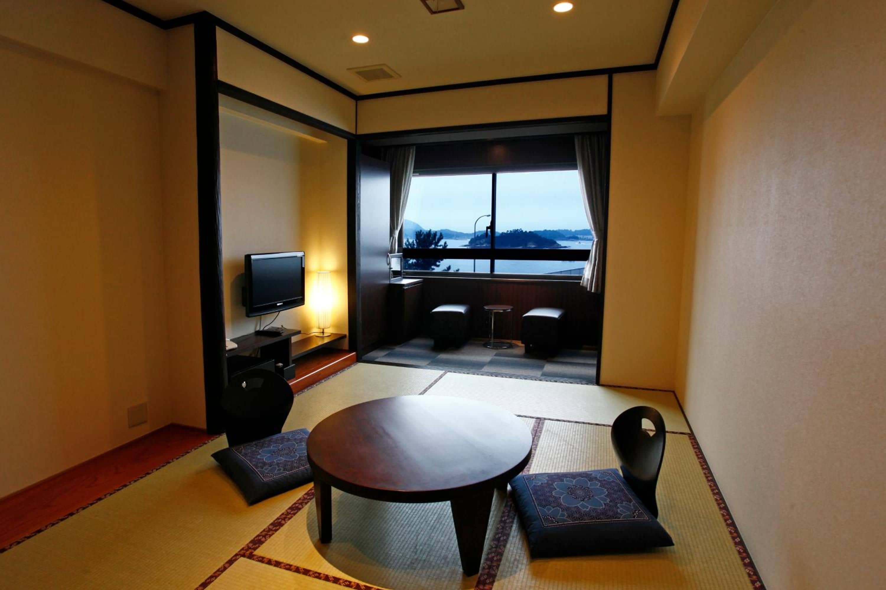A hotel room with tatami flooring, a round table, two cushioned seating, a television, and a view of a body of water.