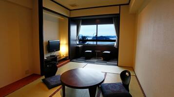 A hotel room with tatami flooring, a round table, two cushioned seating, a television, and a view of a body of water.