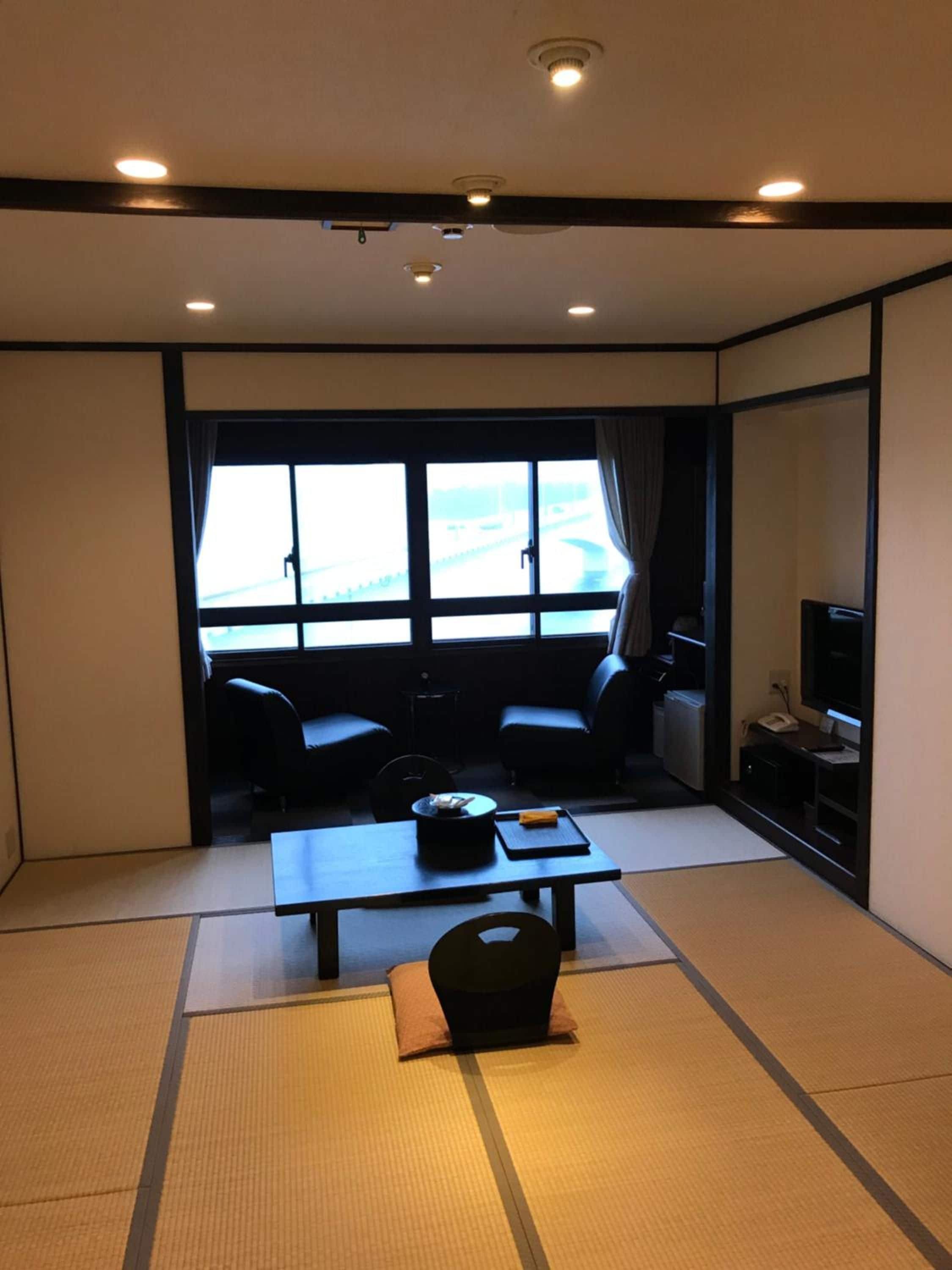 A room with tatami flooring, a low table with a black tray, two black chairs, a television, and a window with curtains.