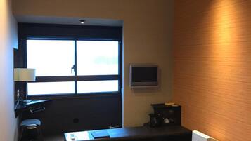 A modern room with a desk, chair, and a small table with cushions. There is a window with blinds, a flat-screen TV mounted on the wall, and a radiator on the floor.
