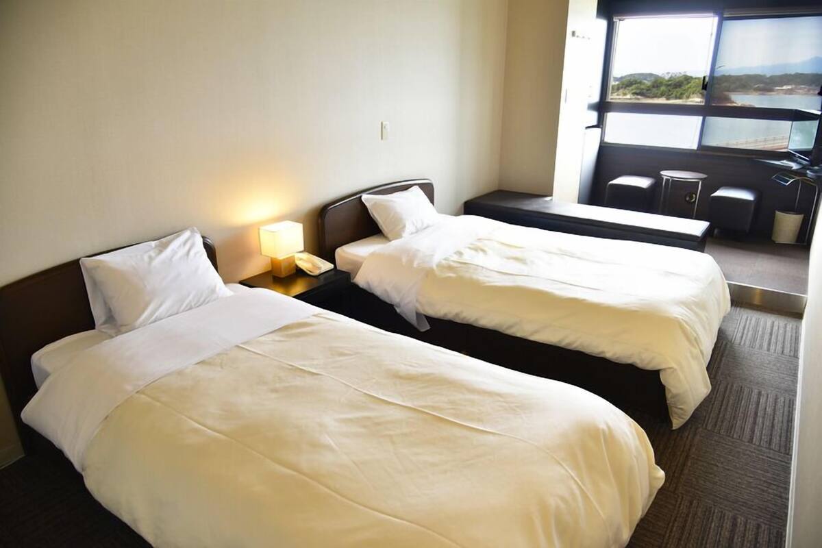 A hotel room with two beds, a bedside table with a lamp, a small table with chairs, and a large window with a view.