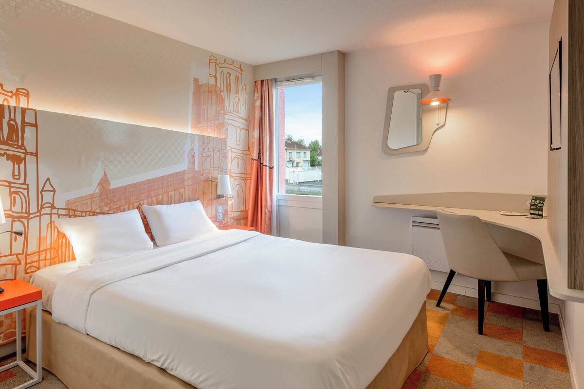 A hotel room with a large bed, a desk, and a chair. There is a window with curtains, a wall-mounted mirror, and a wall with a cityscape mural.