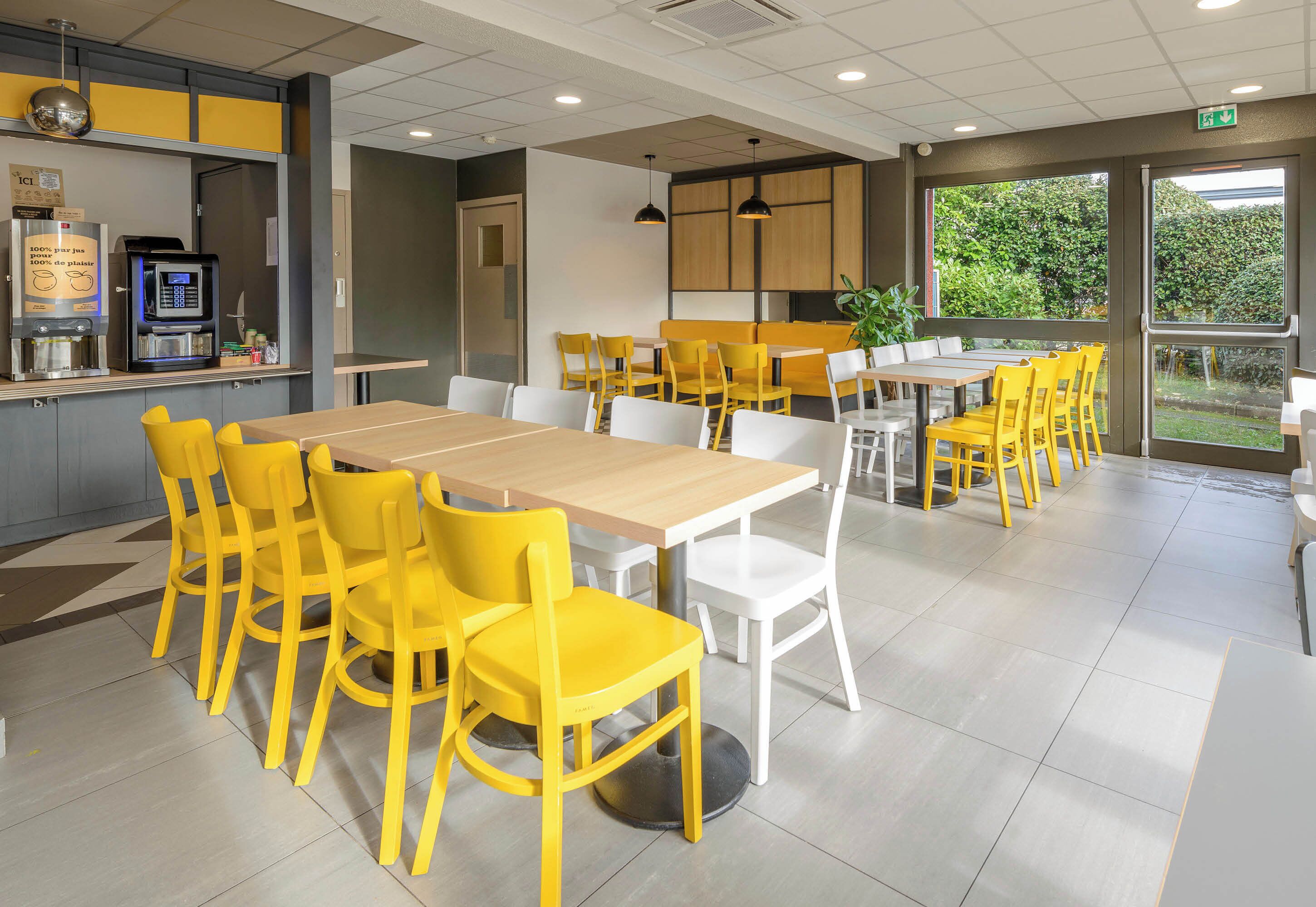 A modern dining area with yellow and white chairs, a coffee machine, and a view of greenery through large windows.