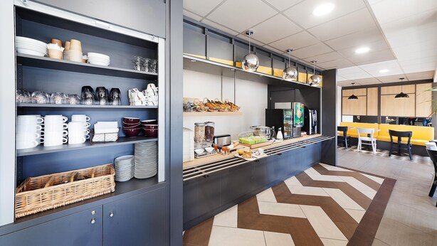 A modern kitchen with built-in shelves for dishes and utensils, a central island with a coffee machine, and a dining area with chairs and a yellow bench. - B&B Hôtel Albi (Albi)