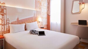 A hotel room with a large bed, a laptop on the bed, a nightstand, a chair, and a wall-mounted mirror. - B&B Hôtel Albi (Albi)
