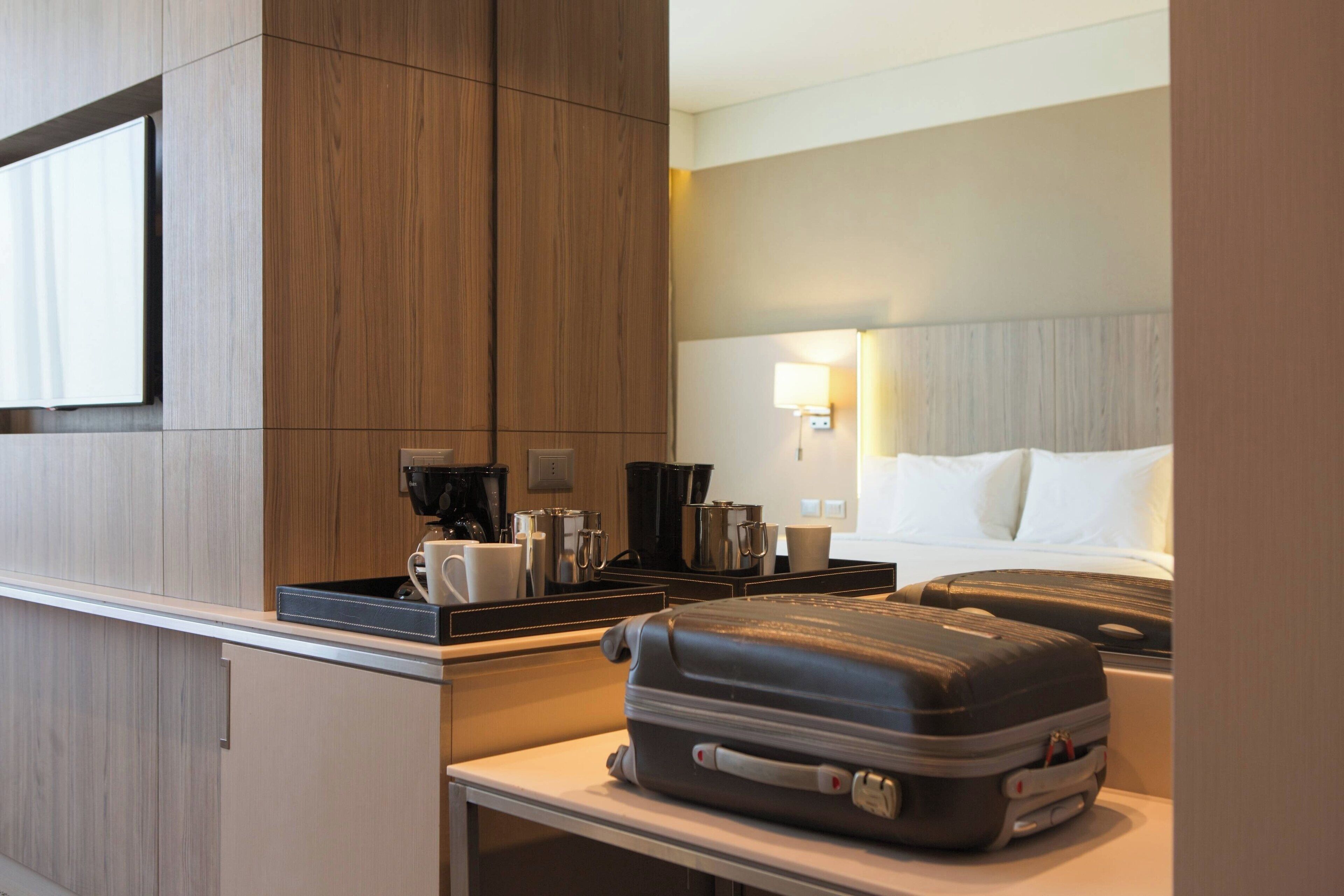Quarto de hotel com uma cama, uma mesa de cabeceira com uma cafeteira e uma mala sobre a mesa.