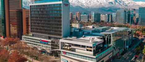 A modern building with a glass facade and a rooftop garden, surrounded by other high-rise buildings and a mountain range in the background.