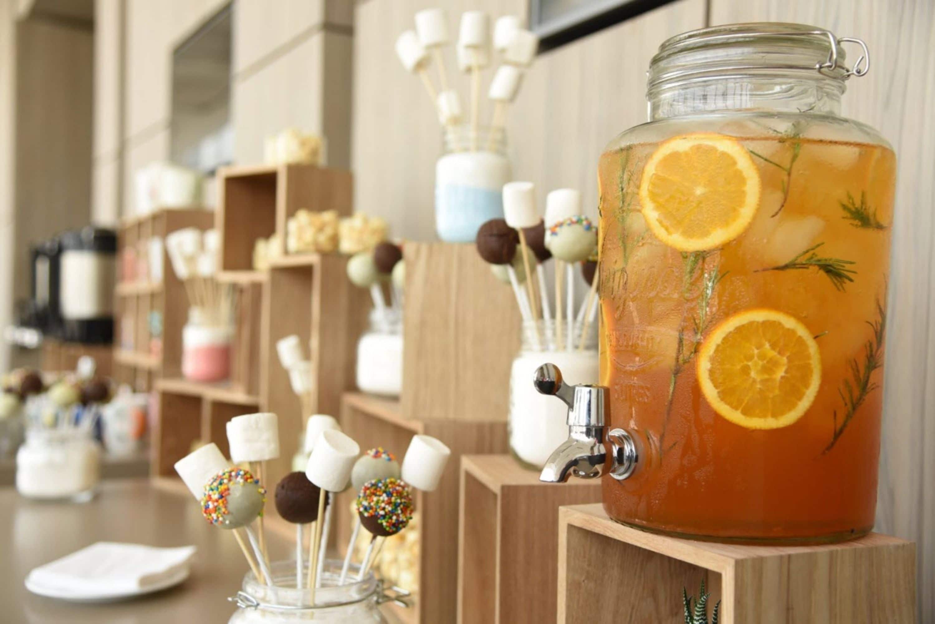 Um distribuidor de bebidas com um jarro de vidro cheio de líquido, decorado com rodelas de laranja e ervas.