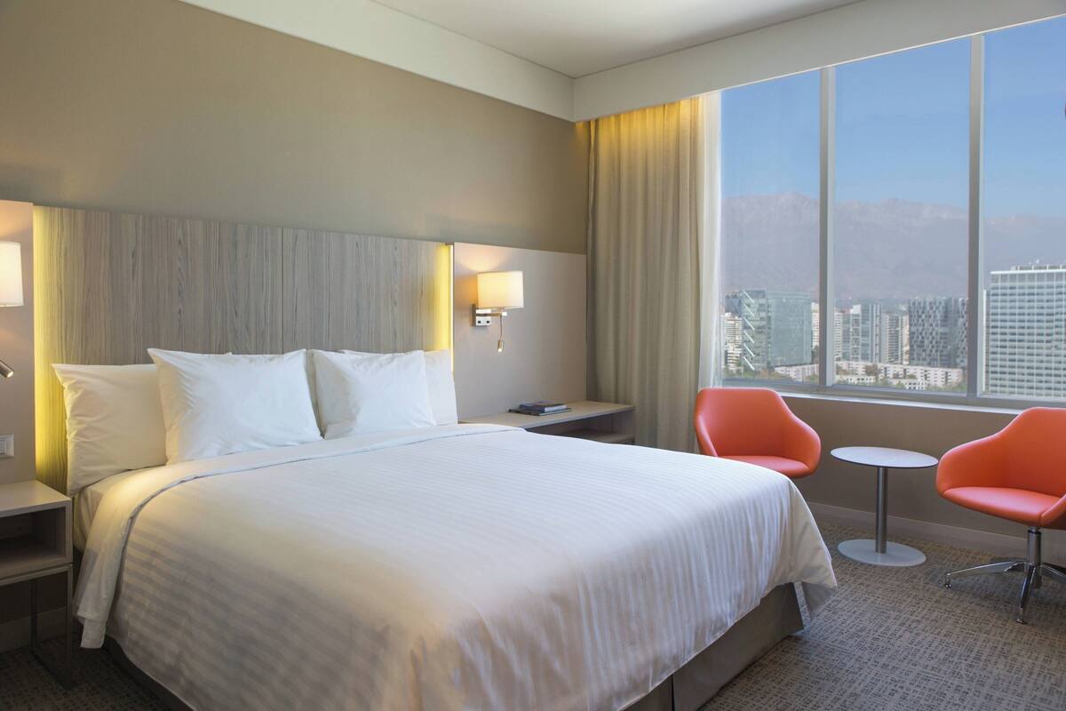 Ein Hotelzimmer mit einem großen Bett, zwei Nachttischen, einem Schreibtisch mit Lampe, zwei orangefarbenen Sesseln und einem Fenster mit Blick auf die Stadt.