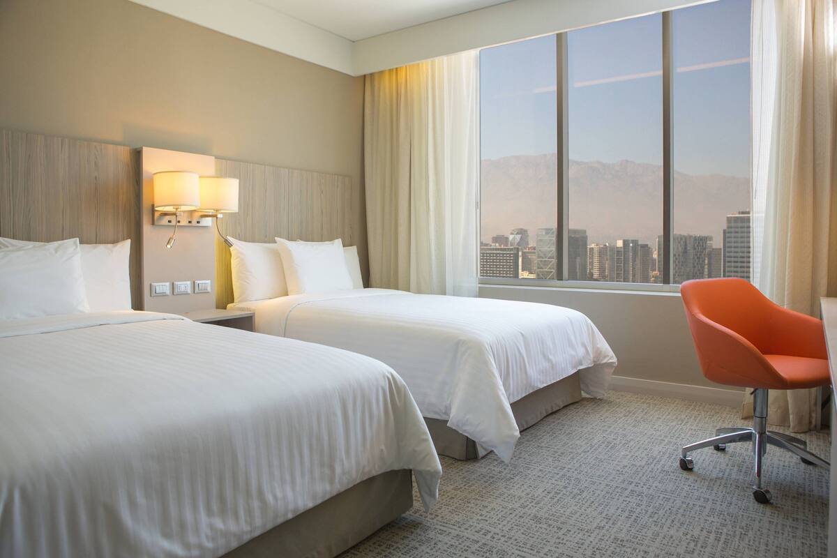 Ein Hotelzimmer mit zwei Betten, einem Schreibtisch, einem Stuhl, einer Lampe und einem großen Fenster mit Blick auf die Stadt.