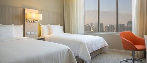 A hotel room with two beds, a desk, a chair, a lamp, and a large window with a city view.