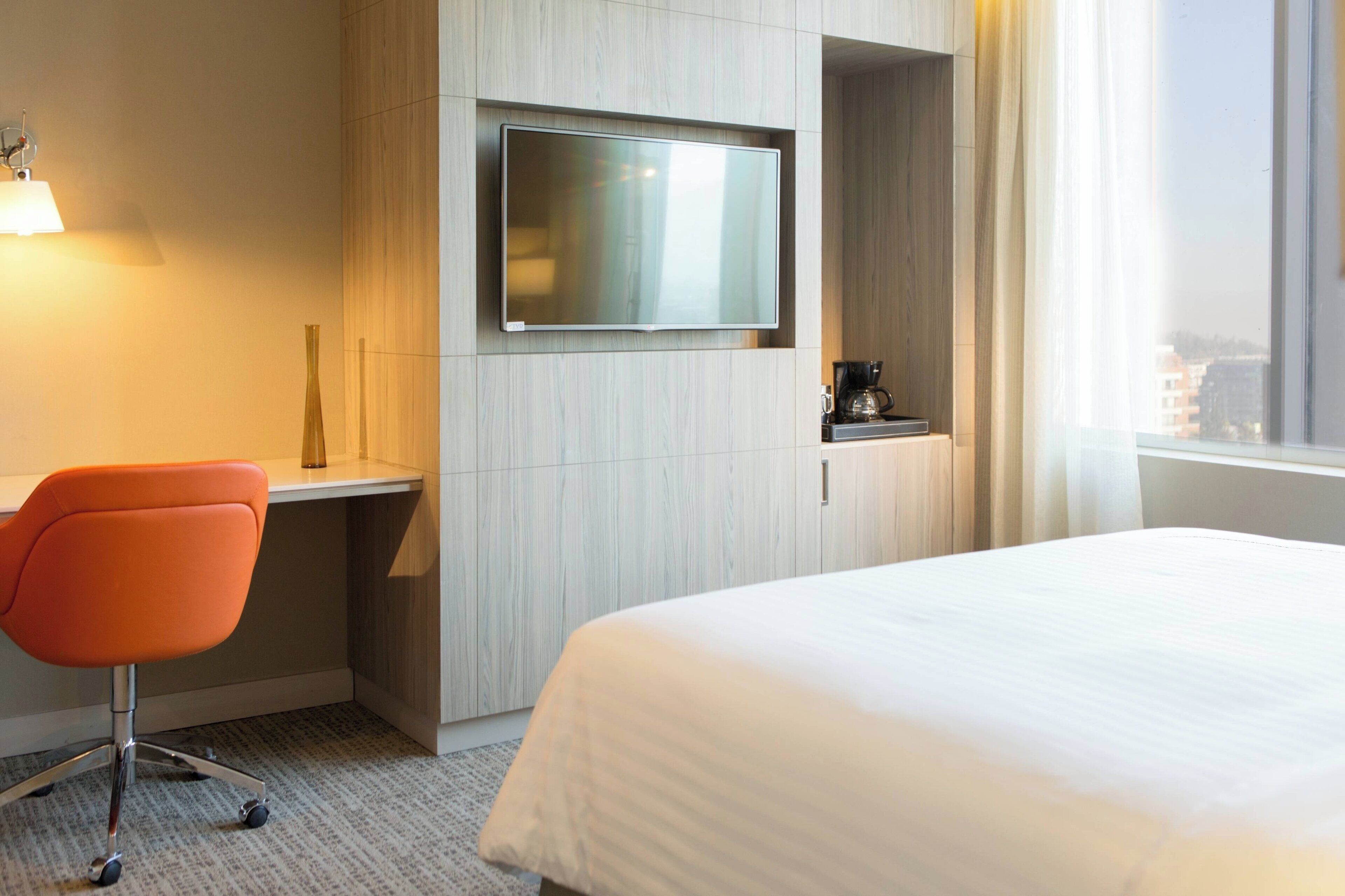 Een hotelkamer met een bed, een bureau, een stoel, een aan de muur bevestigde televisie en een raam met uitzicht op gebouwen.