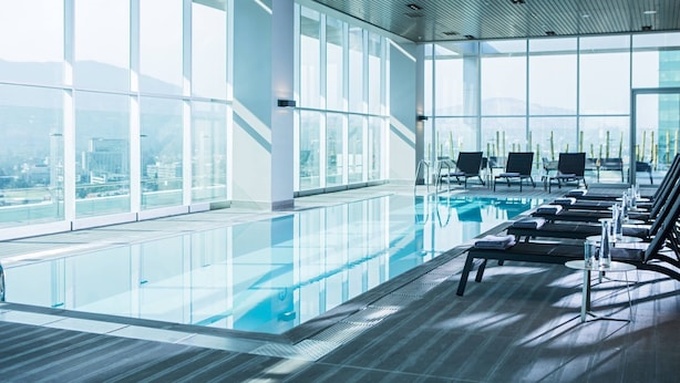 Ein moderner, überdachter Swimmingpool mit Glaswänden, Liegestühlen und Blick auf die Stadt.