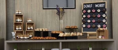 A buffet setup with various food items, including donuts and pastries, displayed on a wooden table against a wall with a mounted television and a sign reading "DONUT WORRY".