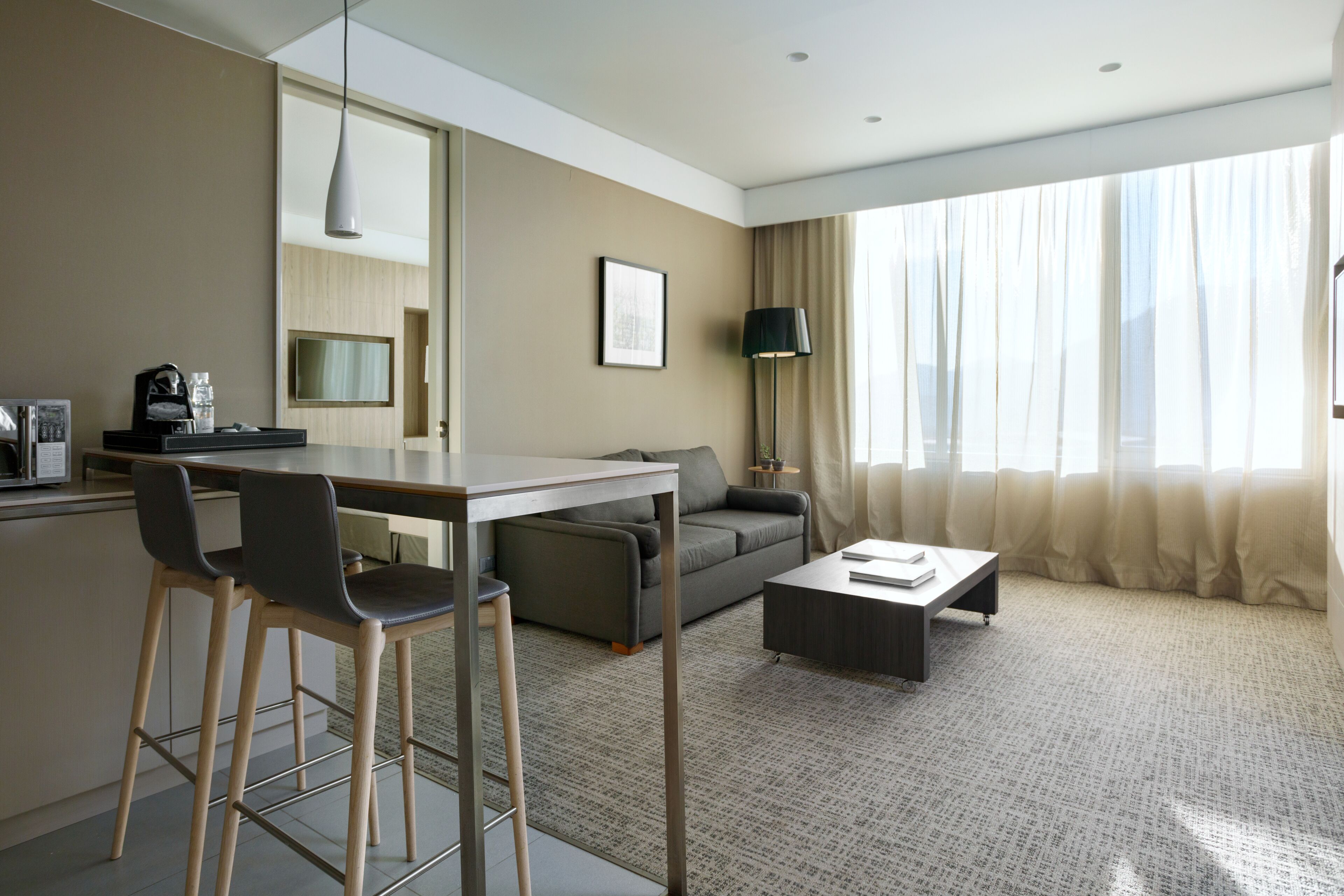 A modern hotel room with a dining area, sofa, and a large window with curtains.
