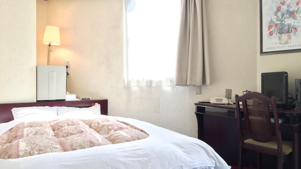 Ein Hotelzimmer mit Bett, Schreibtisch, Stuhl, Lampe und einem fensterbepflanzten Fenster.