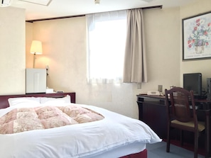 Habitació d'hotel amb llit, escriptori, cadira, làmpada i finestra amb cortines.