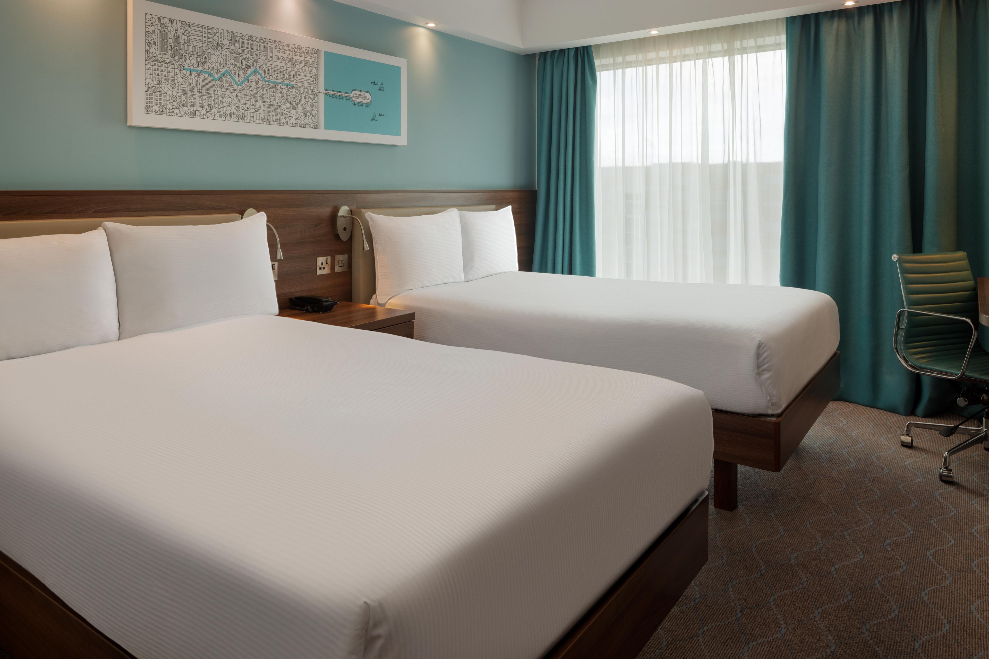 Una habitación de hotel con dos camas, una mesita de noche, una lámpara, una silla y una ventana con cortinas.