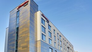 A modern hotel building with a prominent 'Hampton' sign on the corner, featuring a clean, contemporary design with large windows and a clear sky background.