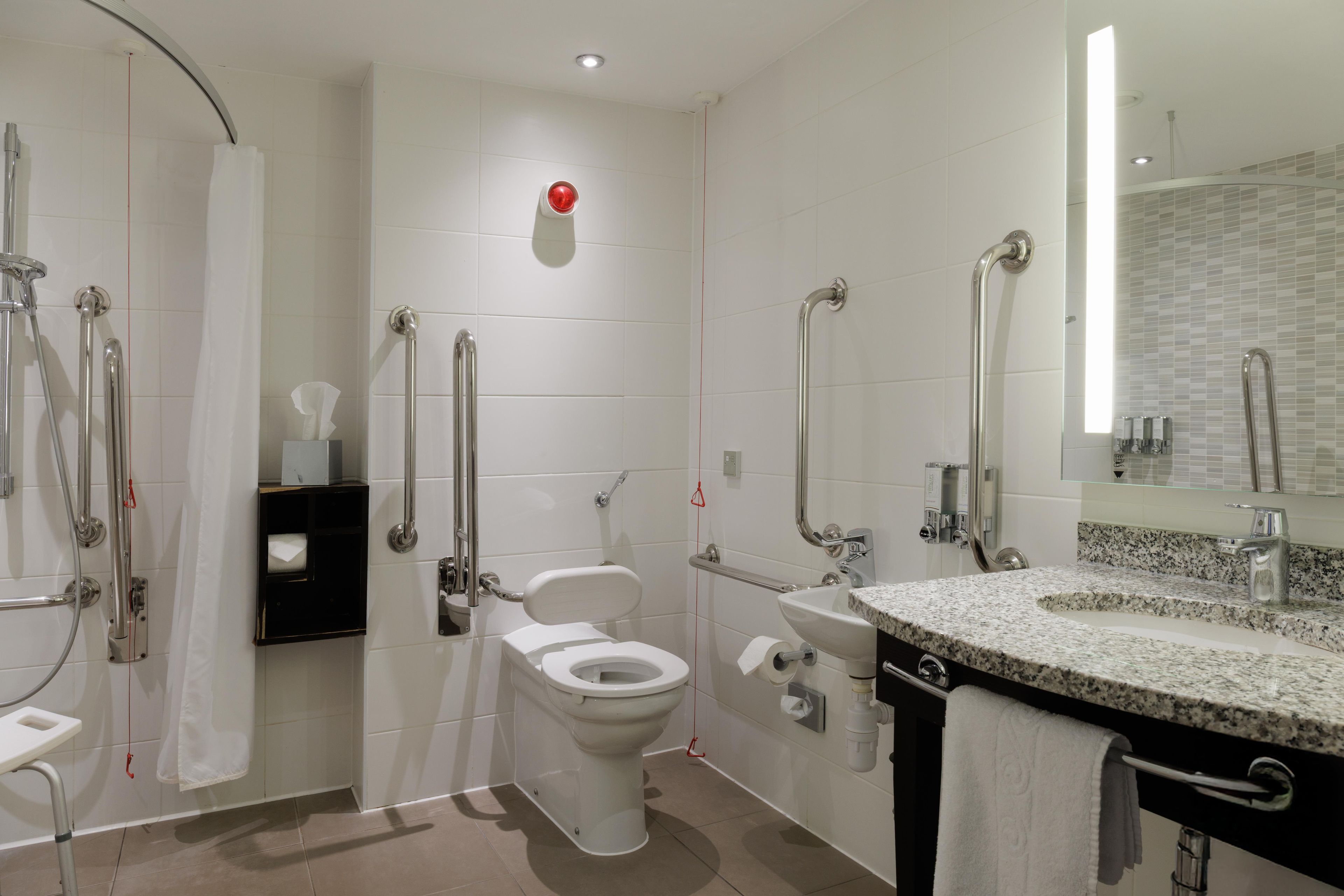 A modern bathroom with a shower, toilet, and sink, equipped with grab bars and a hand dryer.