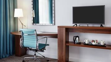A hotel room with a desk, chair, TV, lamp, and a coffee station.