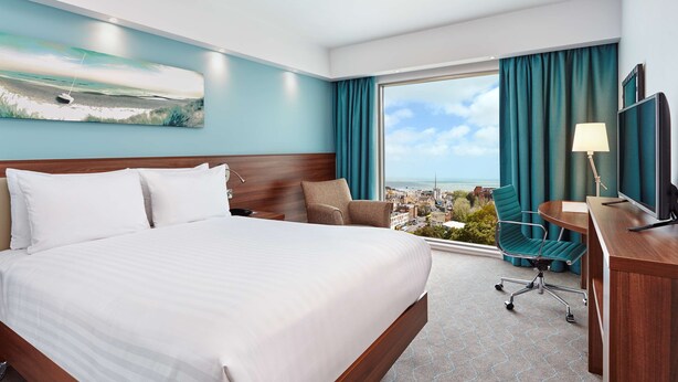 A hotel room with a large bed, a desk with a TV, a chair, and a view of the city through a large window.