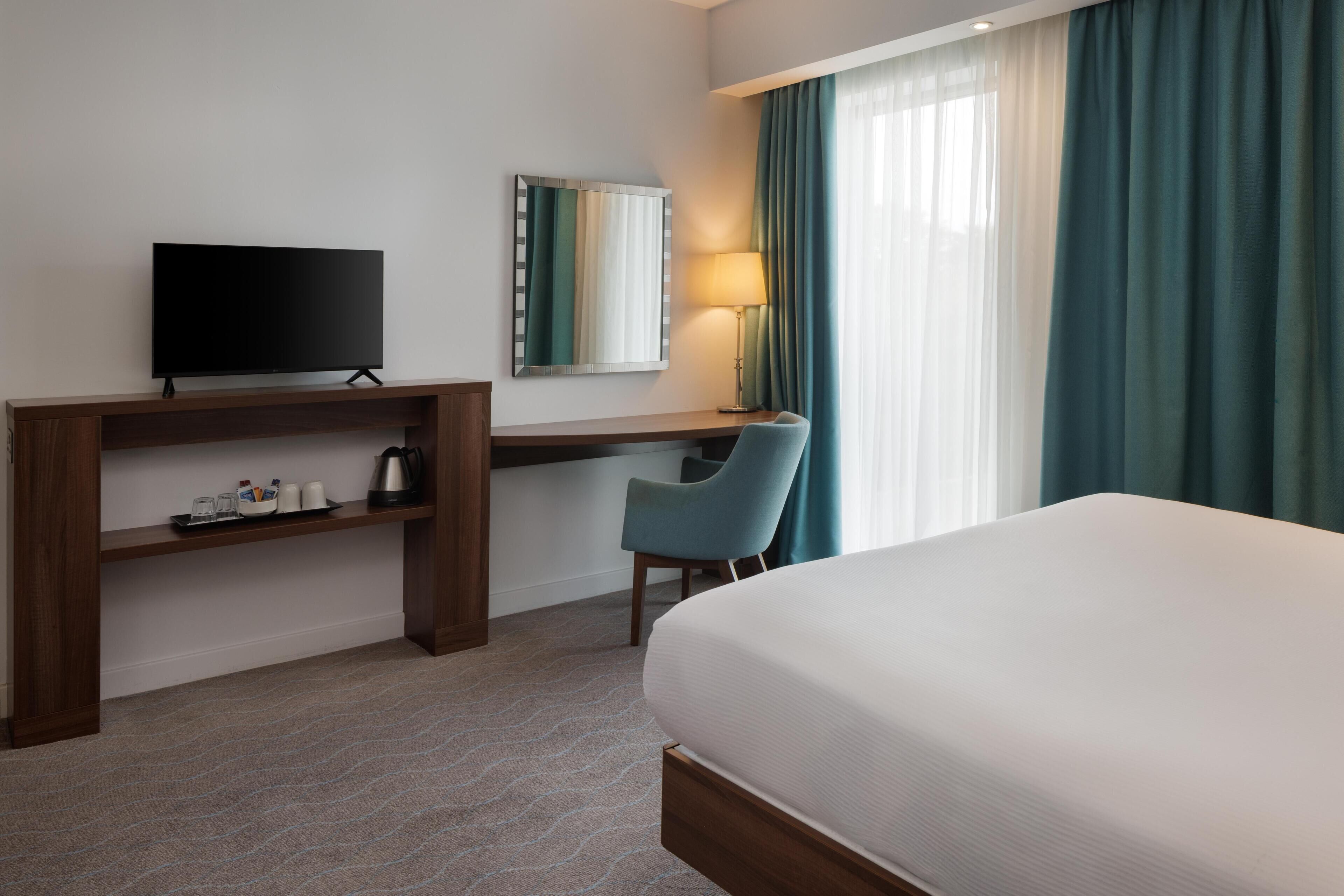 A hotel room with a bed, a desk with a TV, a chair, a lamp, and a window with curtains.