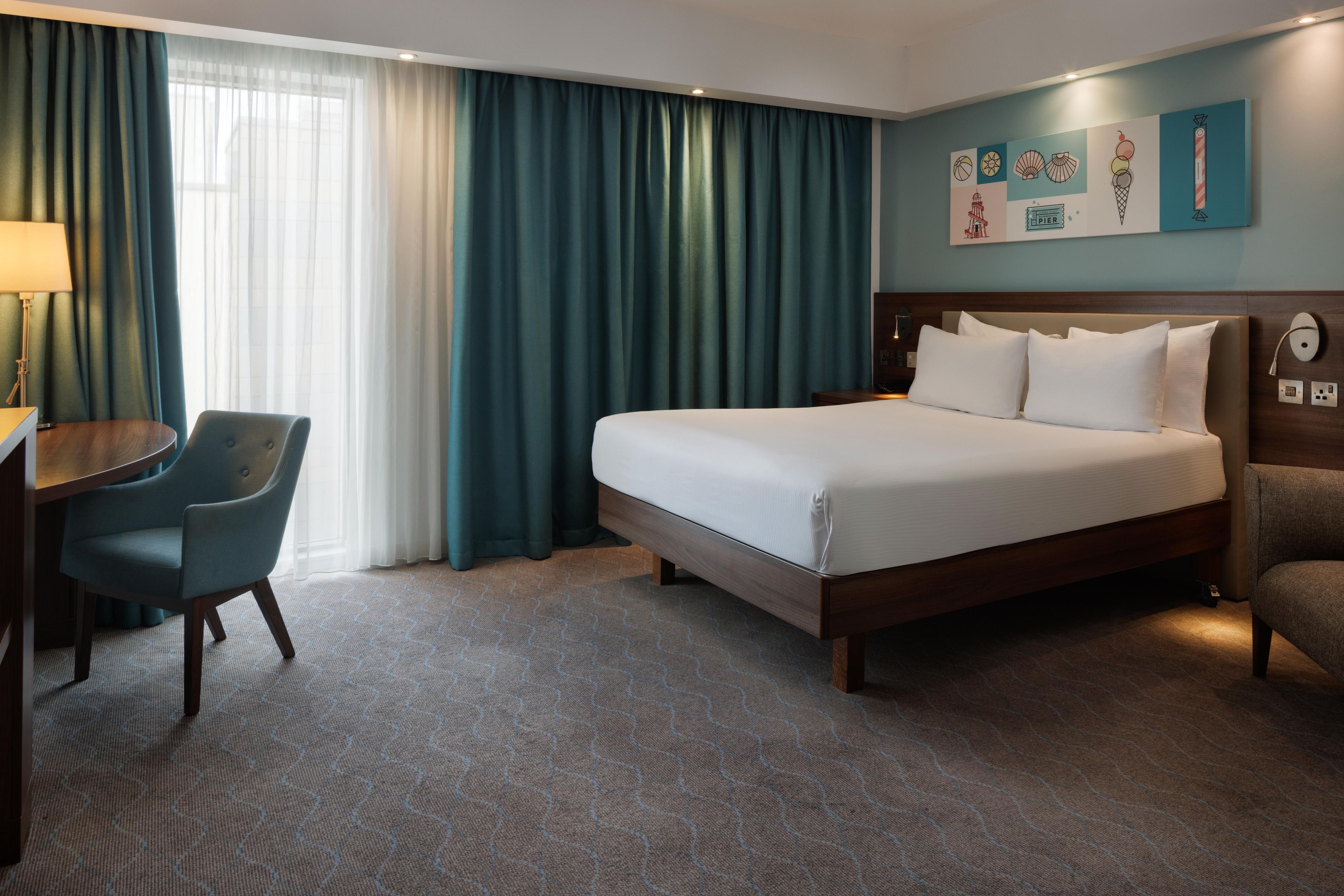 Una habitación de hotel con cama, escritorio, silla y un ventanal con cortinas.