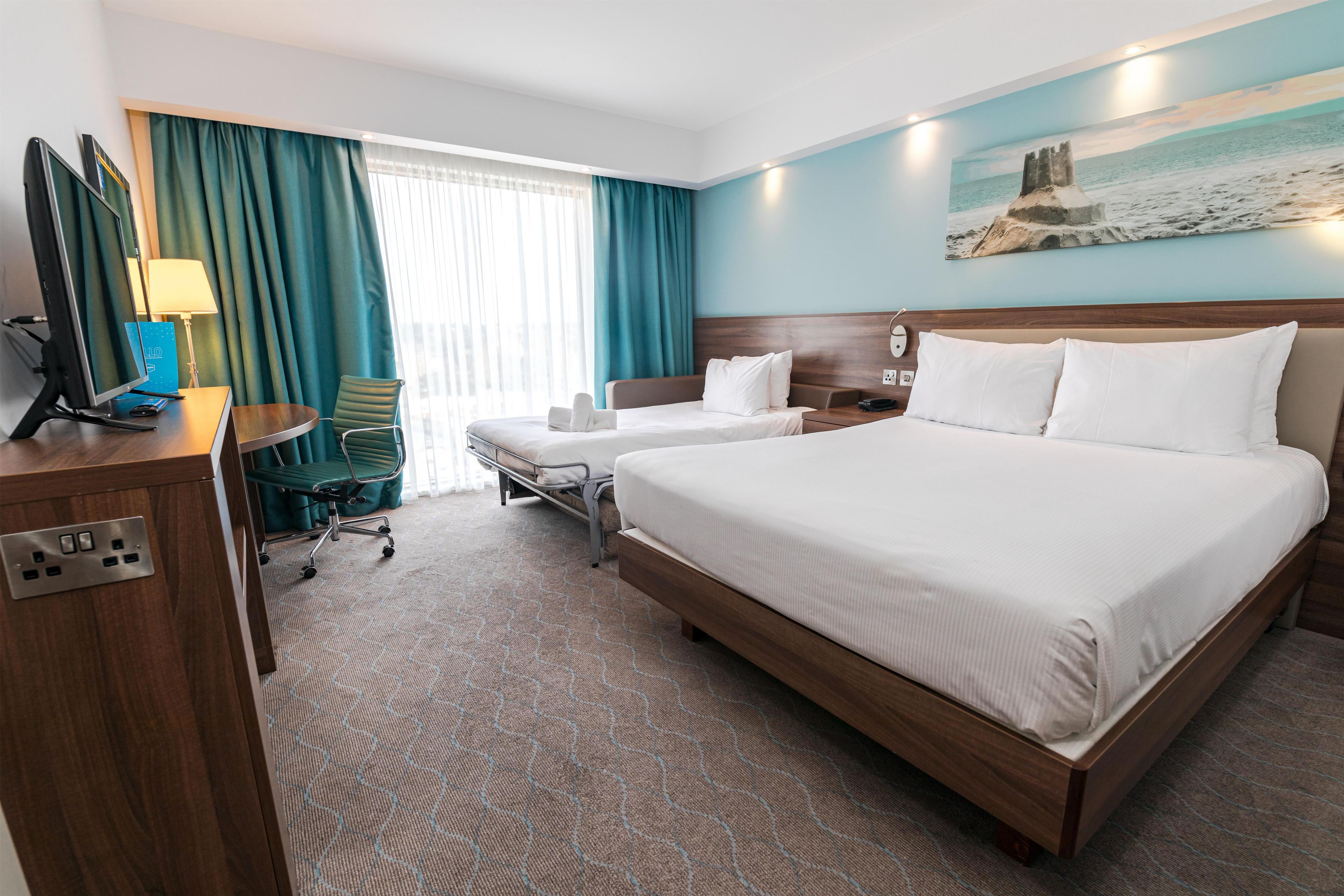 A hotel room with a large bed, a desk with a computer, a chair, and a window with curtains.