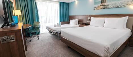 A hotel room with a large bed, a desk with a computer, a chair, and a window with curtains.