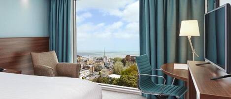 A hotel room with a large bed, a desk with a chair, a TV, and a view of the city and sea through a large window.